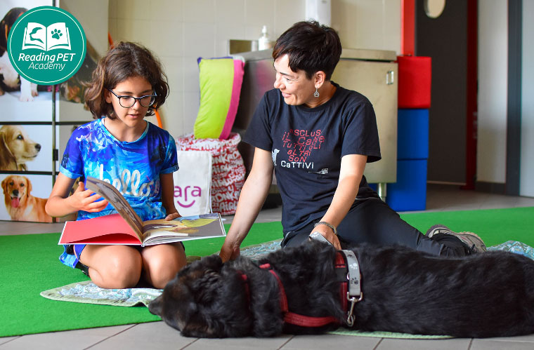 Immagine di una bimba che legge un libro insieme all'insegnante rivolta al cane che le sta sdraiato di fronte